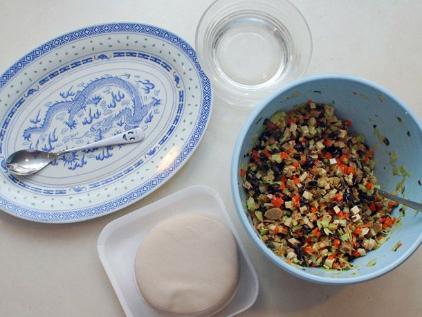 Dumpling wrapping station: plate with spoon, small bowl of water, dumpling wrappers, and a bowl of filling with a spoon. 