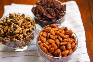 Three glass dishes of spiced nuts on a kitchen towel on a table. The flavors are smoky candied almonds, Mexican spiced chocolate pecans, and olive-rosemary cashews.