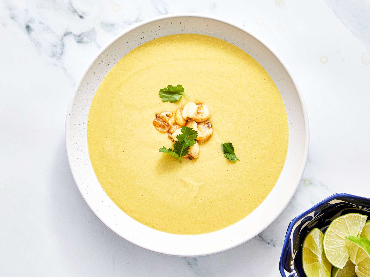 A bowl of corn soup garnished with cilantro and sauted corn kernels lime wedges in a dish beside