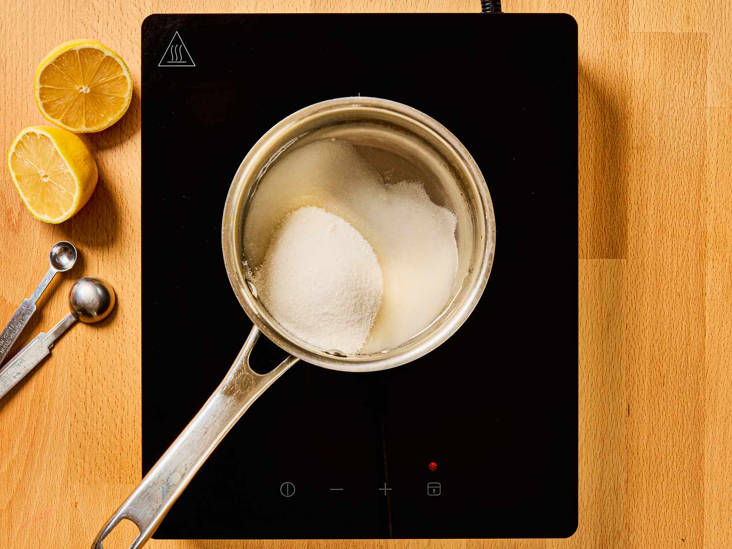 Sugar, water and lemon juice cooking in small pot, with measuring cups and halved lemon on the side