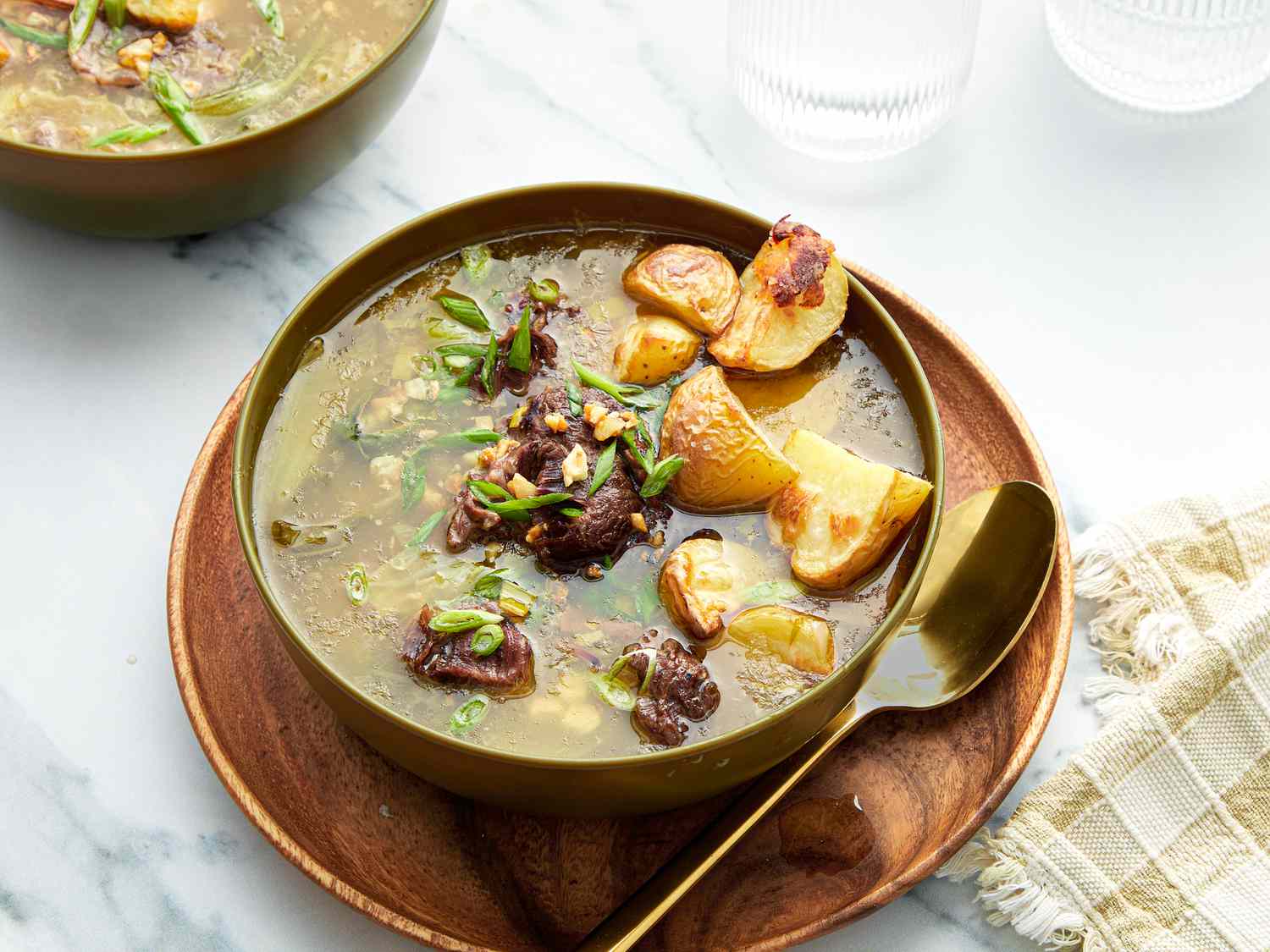 Nilagang Beef on white marble surface with gold spoon. Side of water glass, fabric napkin, and larger bowl on the sides. 