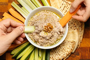 Two hands holding slices of vegetable dipping into a bowl of walnut skordalia surrounded by flatbread and vegetable sticks