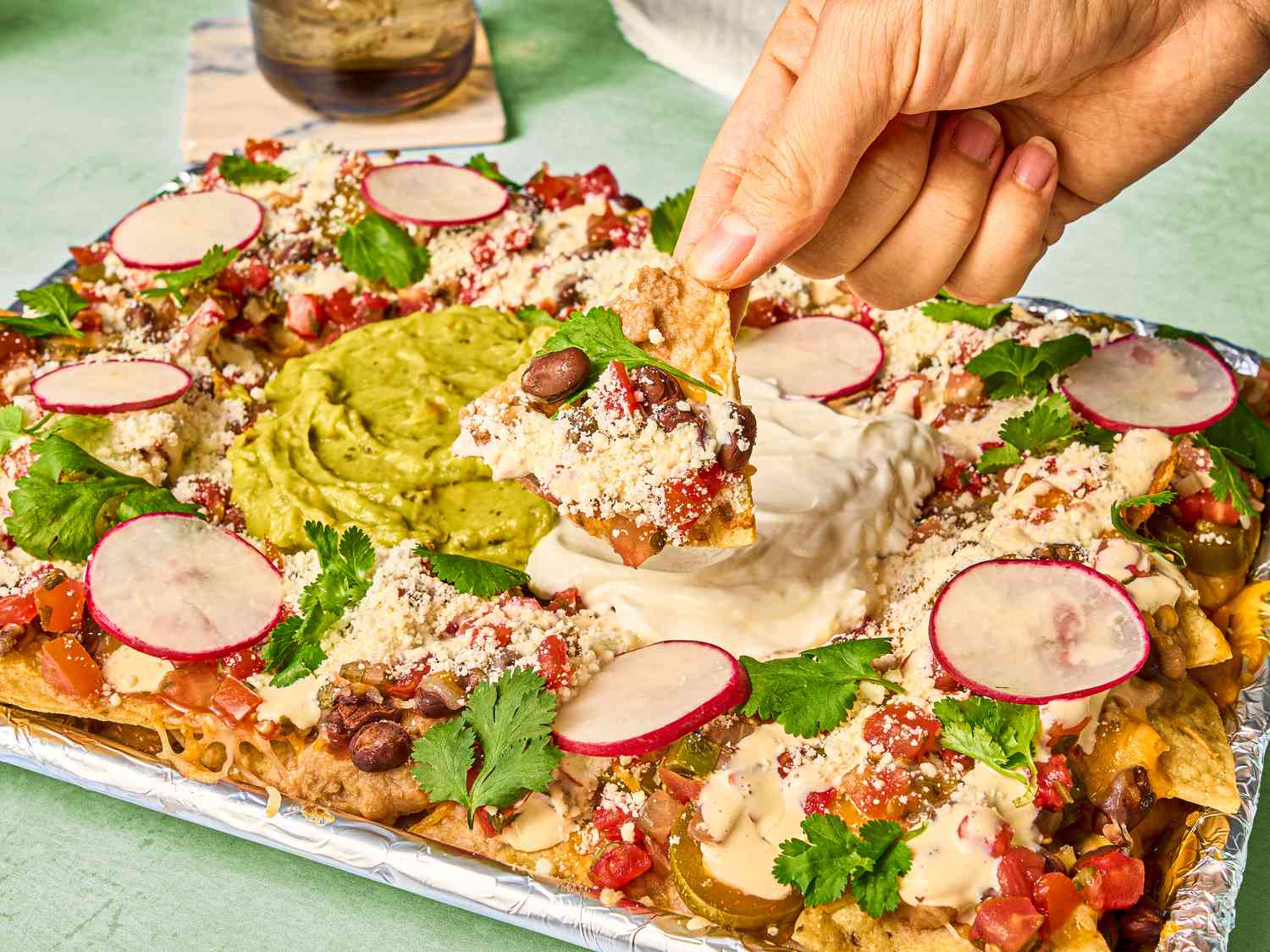 Nachos in foil, with a hand dipping a chip into toppings on a green surface, with a drink in the background