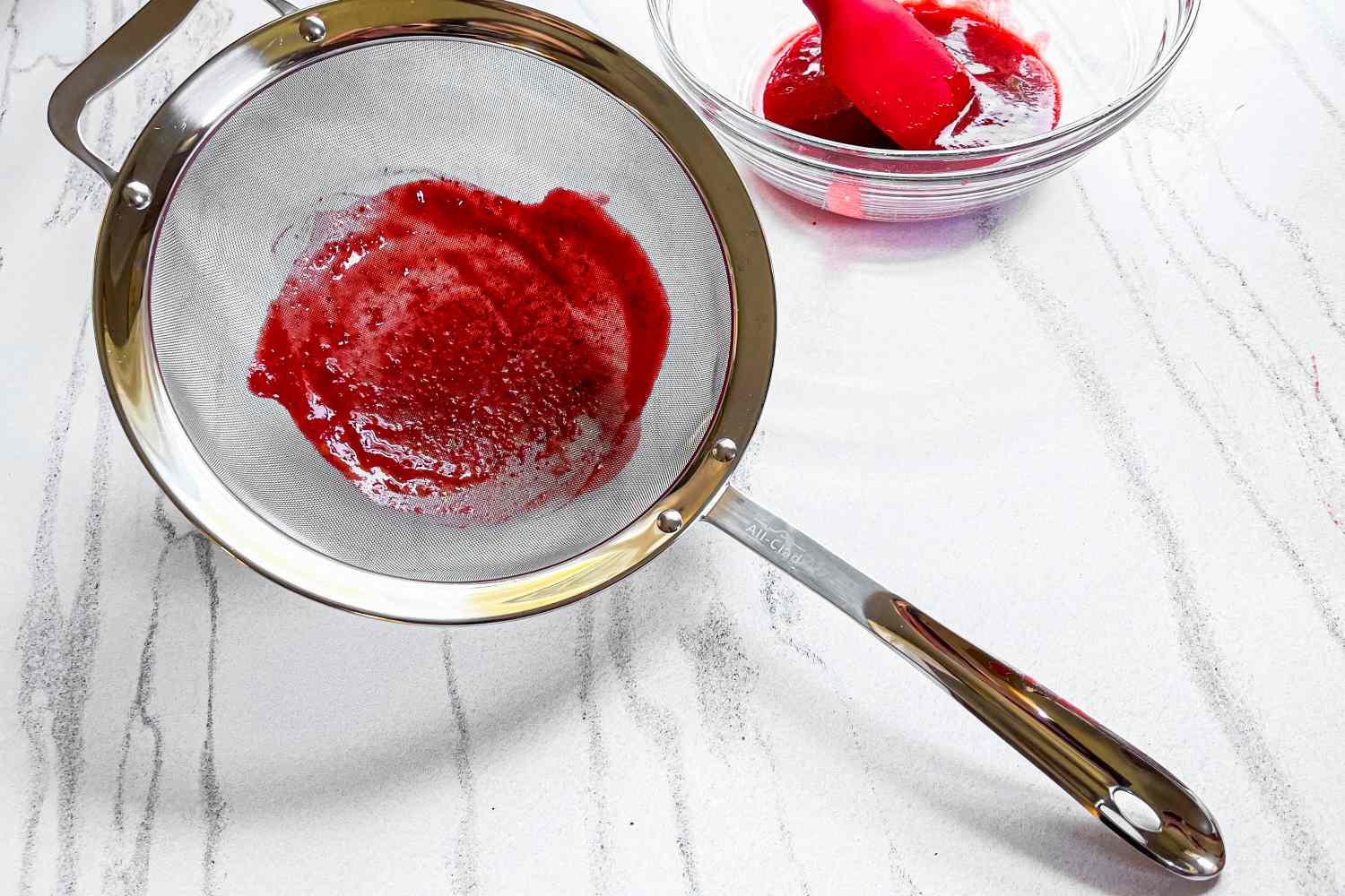 All-Clad Stainless-Steel Strainer, Set of 3 with red puree alongside a bowl with similar contents and a spatula on a surface