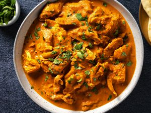 Chicken tikka masala in a white speckled ceramic bowl. In the top left corner is a small bowl holding cilantro leaves and on the top right edge of the image is a plate holding naan bread.