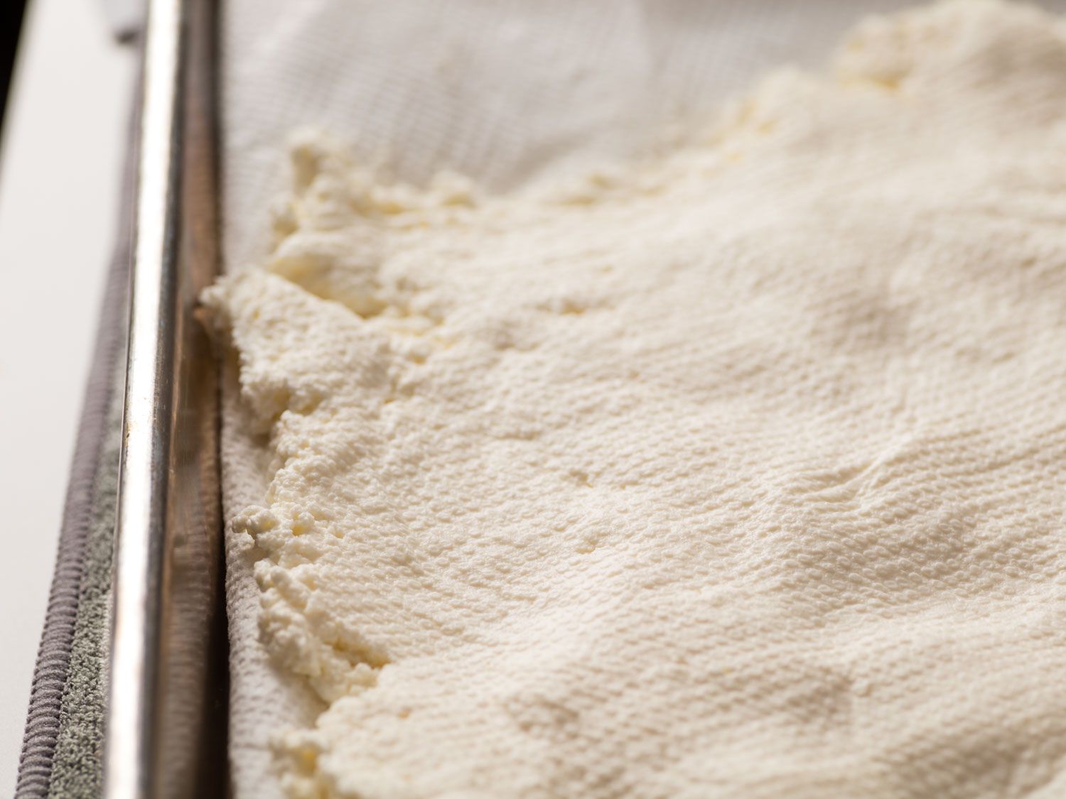 Close-up of a paper-towel-lined sheet pan with ricotta spread thinly over it. The surface of the ricotta is imprinted with the texture of the paper towel that was used to dry the top.