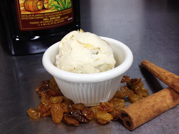 A scoop of rum raisin ice cream served in a ramekin. A bottle of dark rum, golden raisins, and cinnamon sticks are nearby.