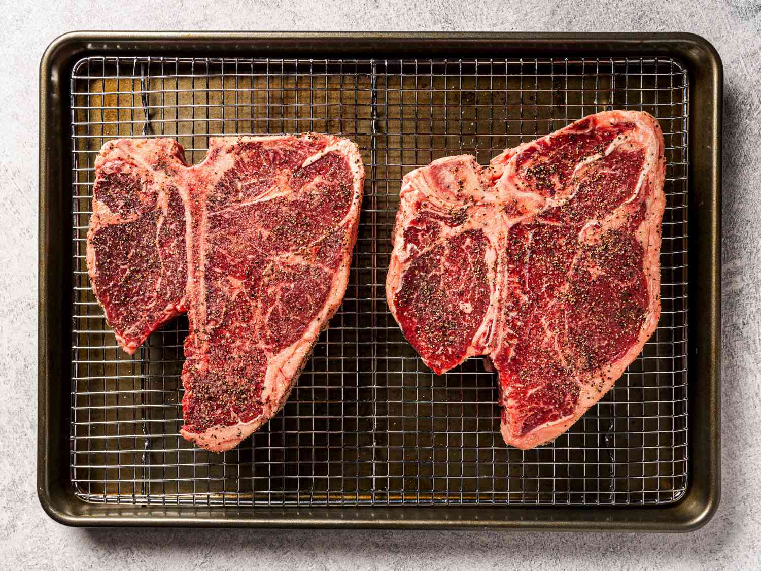 Two prime-grade t-bone (porterhouse) steaks placed on a metal rack set inside of a quarter sheet pan. The steaks have been heavily seasoned with salt and pepper and this shot shows off both the seasoning and the color and marbling of the steaks.