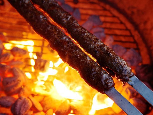 Two Adana kebab skewers held high above a flaring charcoal fire.