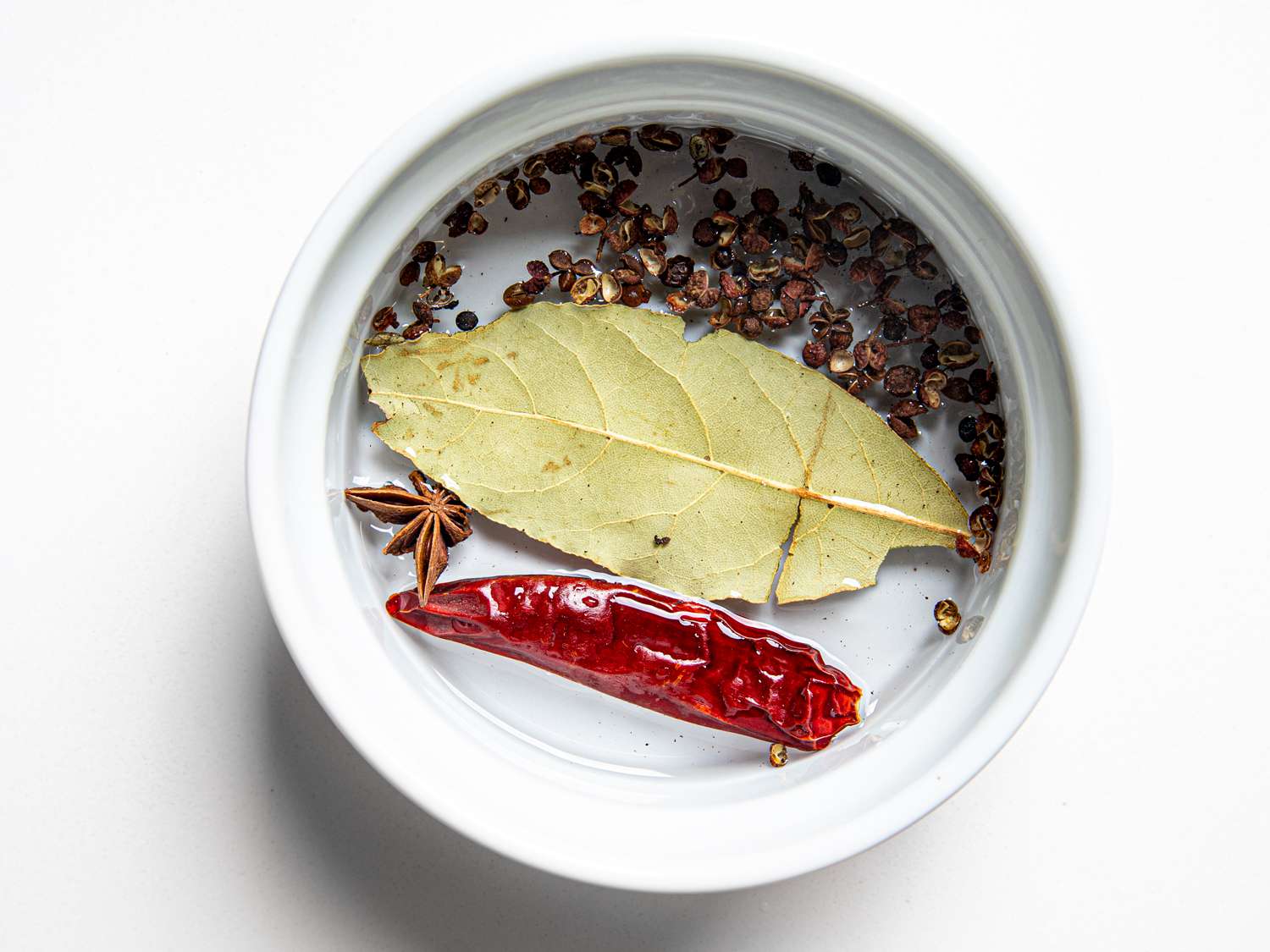 Bay leaf, Sichuan pepper, dried chile, and star anise are submerged in water in a small bowl