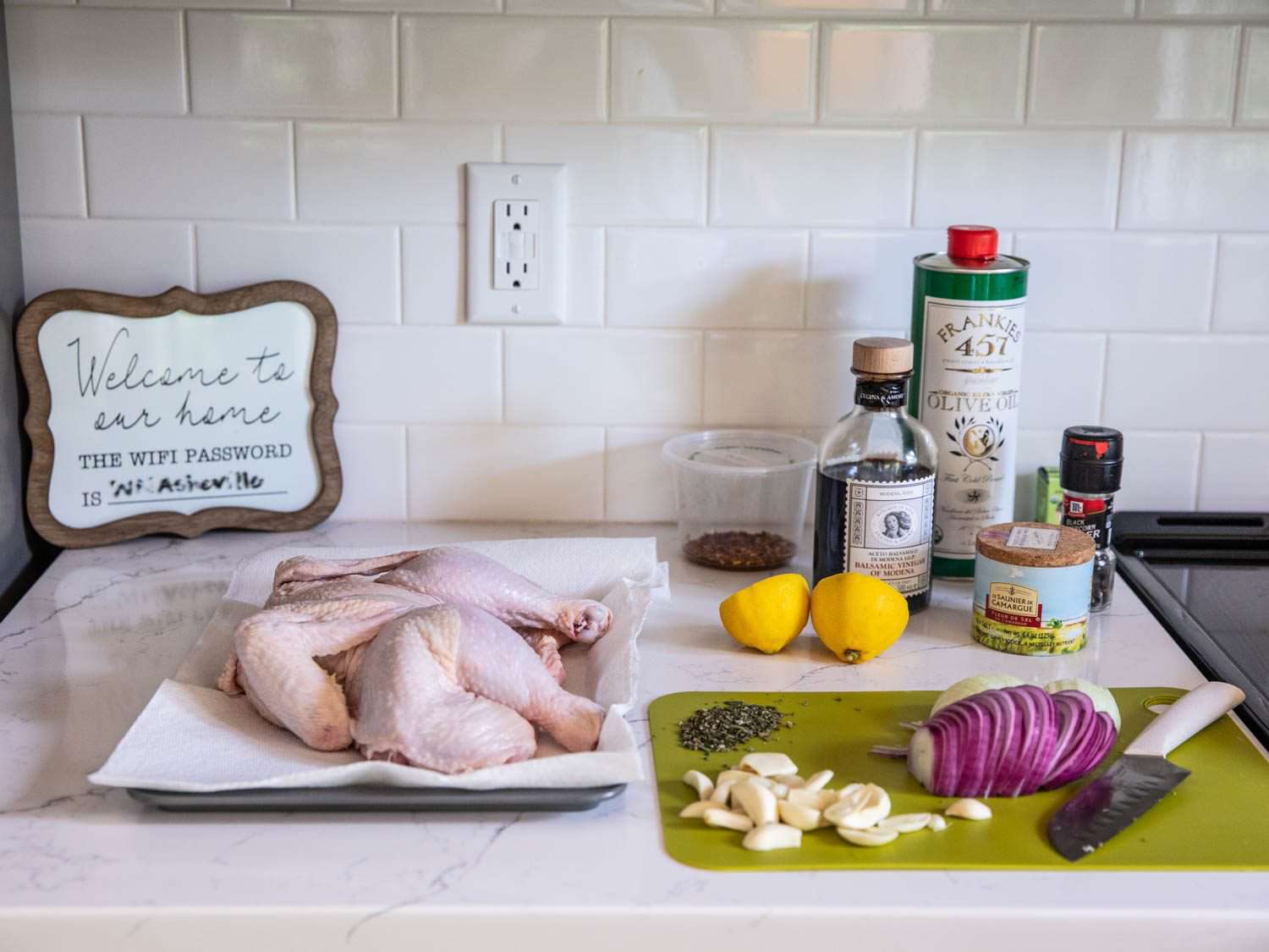 A spread of ingredients on a marble countertop in an Airbnb kitchen: chicken parts on a paper towel–lined tray; onion, garlic, and herbs on a cutting board; balsamic vinegar; fleur de sel; olive oil; grinder of black pepper; lemon halves.