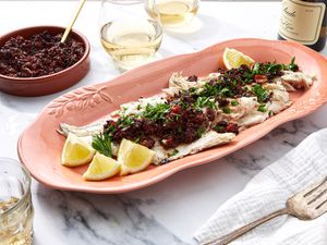 A dish of grilled fish topped with olive and tomato compote served on a table with lemon wedges and parsley garnish