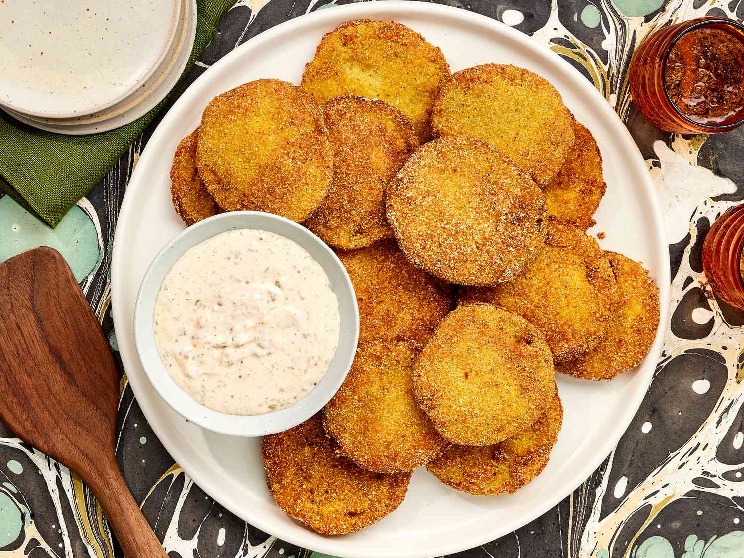Overhead of Horizonal overhead of Fried Green Tomatoes and Rémoulade dip on a printed surface. With Stacked plates, an wooden spoon on the side.