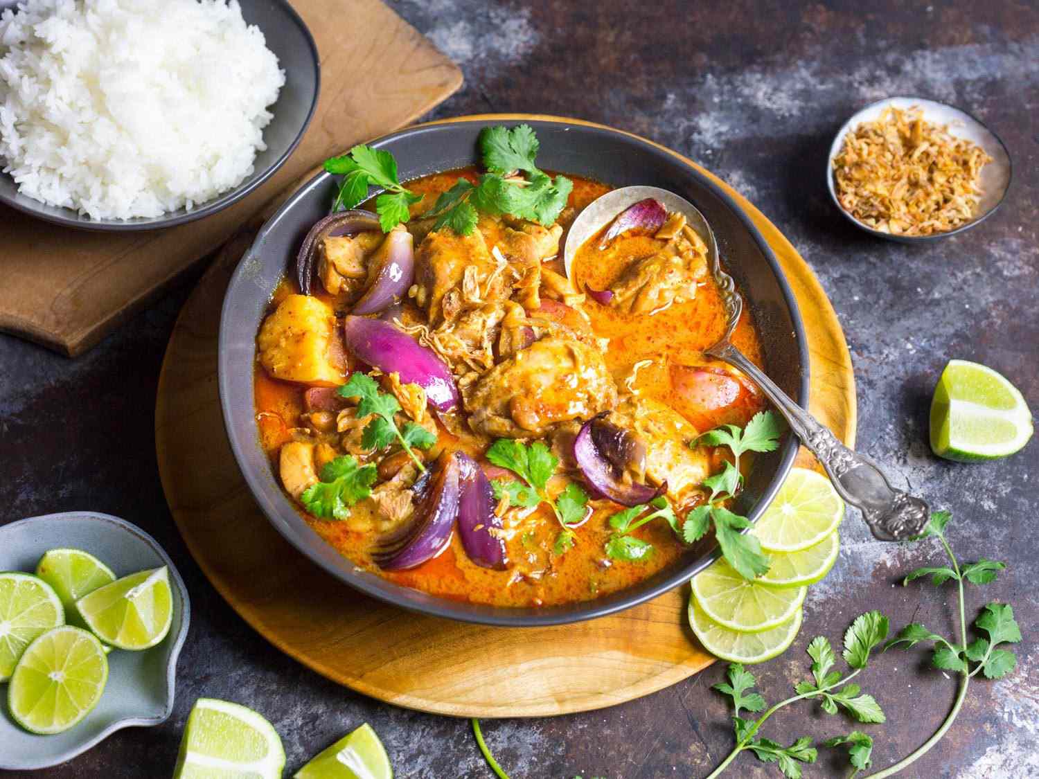 A bowl of chicken massaman curry accompanied with a bowl of rice, lime wedges, and fresh cilantro sprigs.