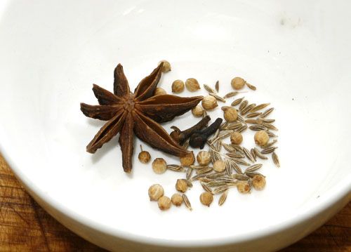 Star anise, whole cumin, whole cloves, and whole coriander seeds in a white bowl.