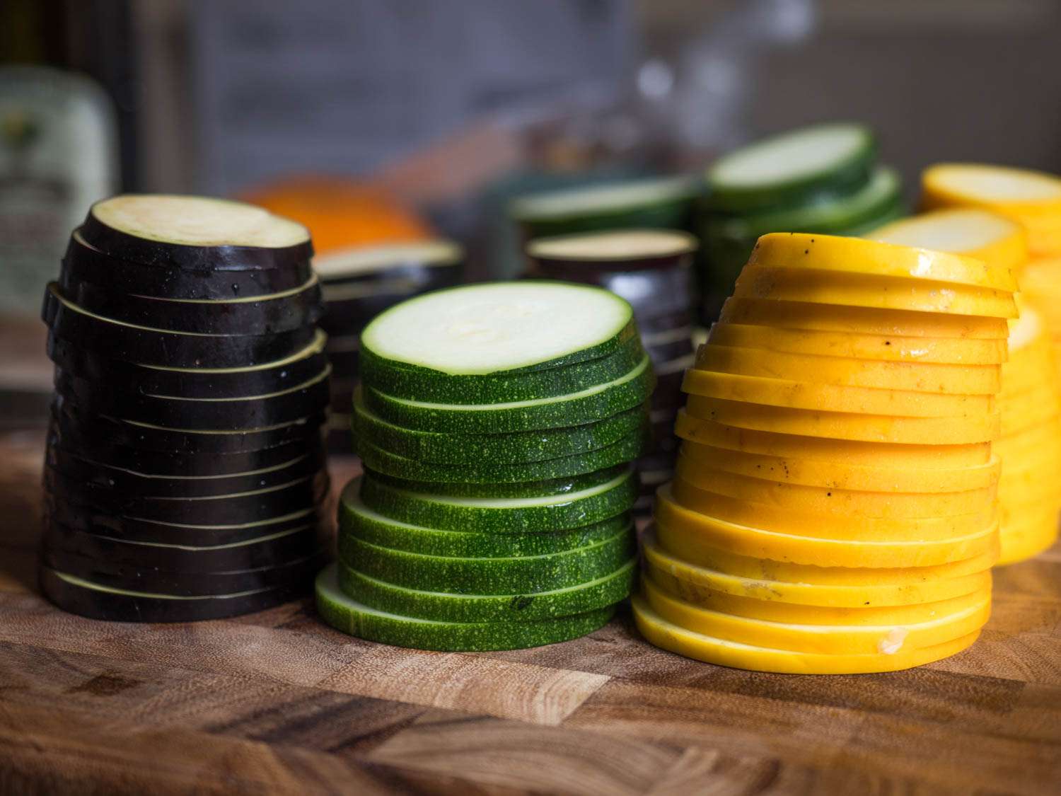 Uniform slices of eggplant, zucchini, and yellow summer squash, stacked so that they're in the same shape they were before being cut.