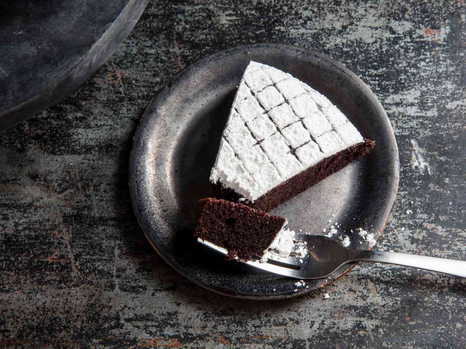 Overhead closeup of a slice of dairy-free chocolate cake served on a small black ceramic plate. A fork cradling a bite of cake rests on the rim of the plate.