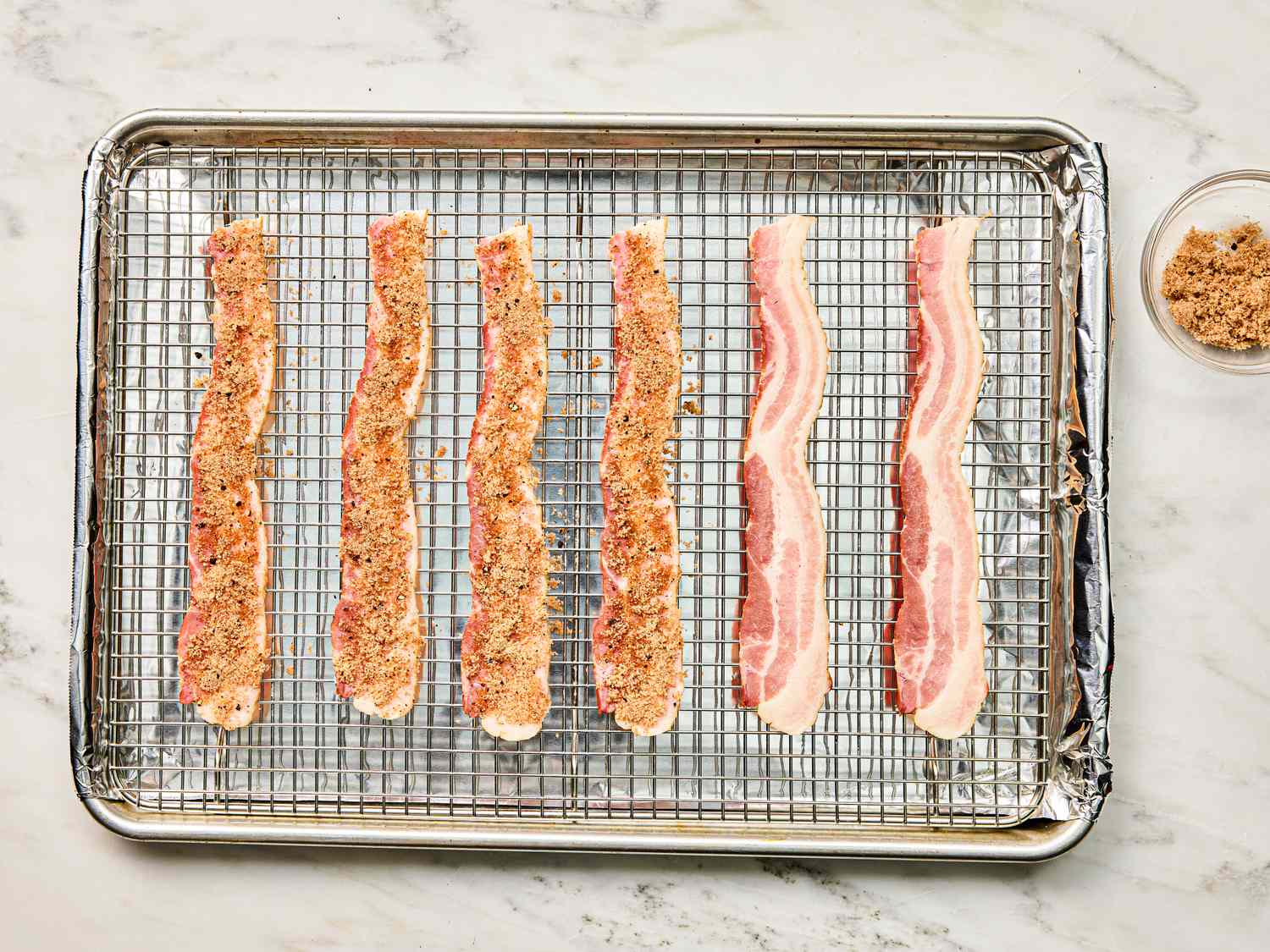 Six bacon strips arranged on a wire rack over a baking sheet with seasoning visibly applied on some strips small bowl of seasoning aside