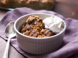 A white ramekin filled with apple crisp and whipped cream, next to spoon on a lavender cloth napkin.
