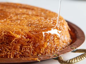 Knafeh being drizzled with syrup from above set on a round copper tray with handles
