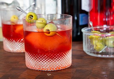 A glass of a cocktail garnished with green olives on a pick placed on a wooden surface with bottles and a dish of olives in the background