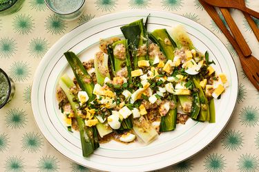 A plated dish of leeks topped with vinaigrette and chopped ingredients, displayed on a patterned surface with tableware