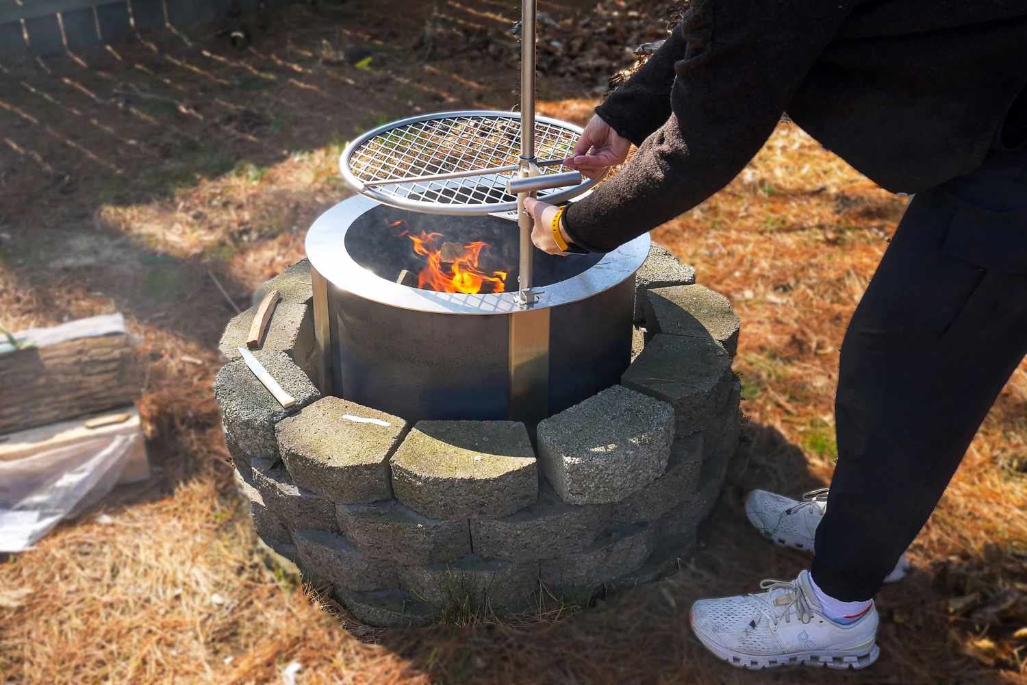 A person adjusting the grill over a fire pit with a burning flame inside