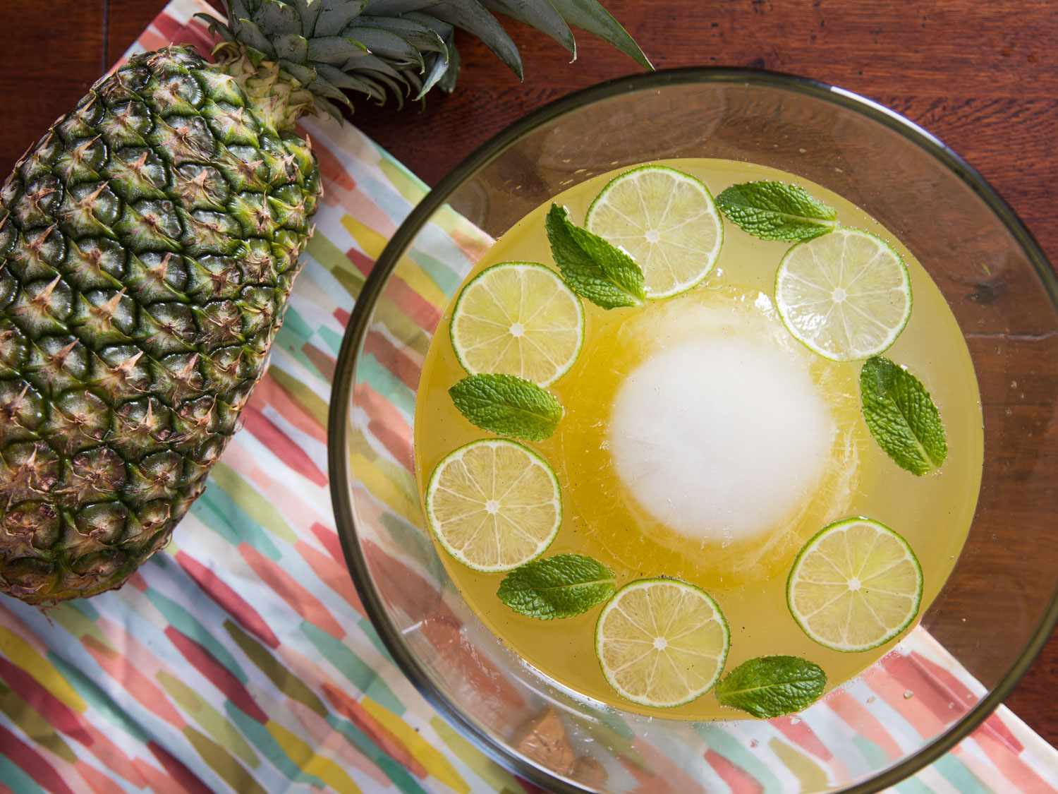 Overhead shot of a glass bowl of sparkling pineapple-rum punch with ring of alternating mint leaves and lime slices and large ball of ice in the center.