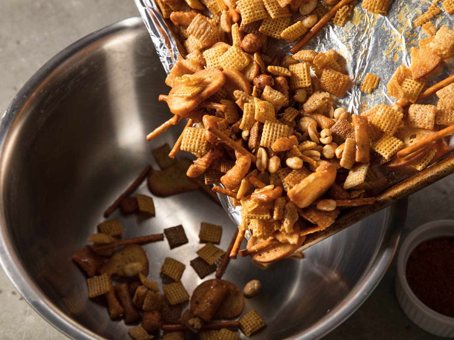 Toasted chex mix being poured from a sheet pan into a metal bowl.