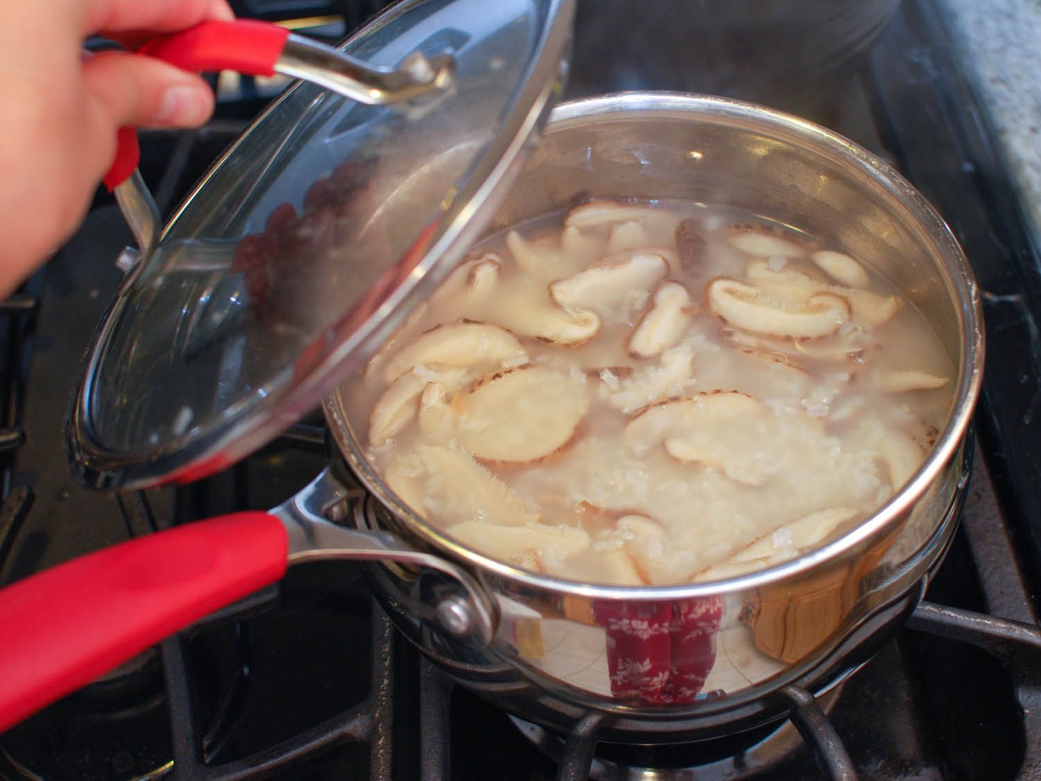 A hand lifting the lid of the congee pot with sliced shiitake mushrooms.