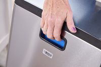 A person lifts the simplehuman 38-Liter Brushed Stainless Steel Steel Trash Can with Lid by the handles