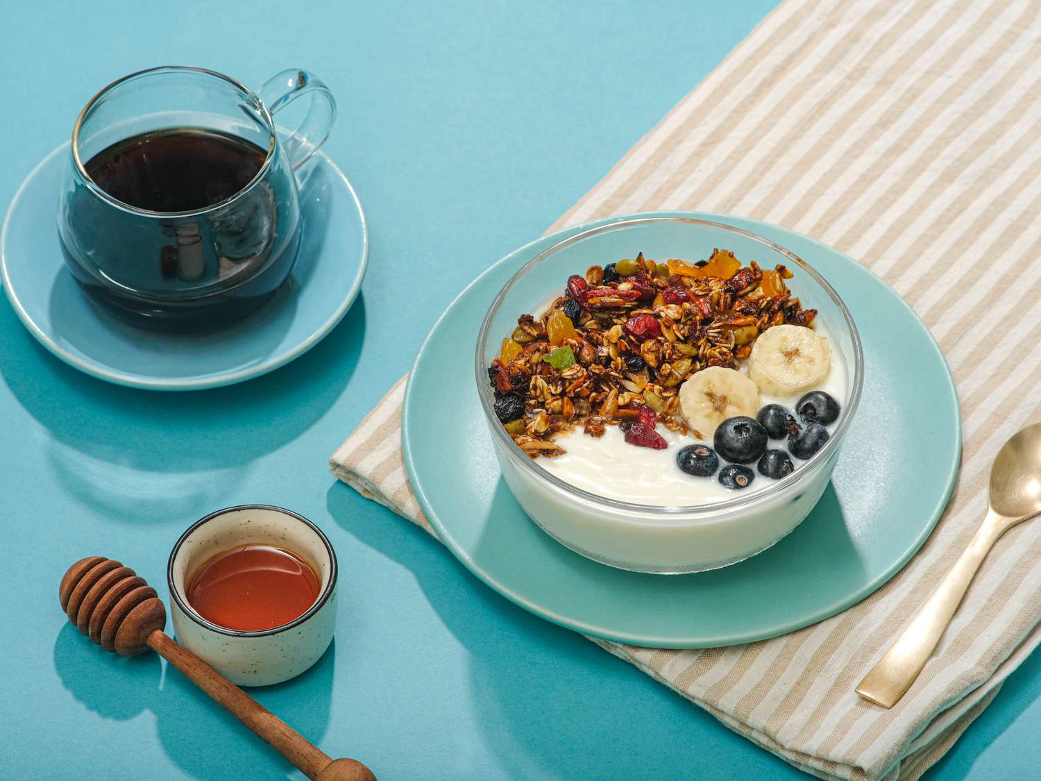 Granola, yogurt, berries, and banana in a glass bowl, on a blue plate, with a striped napkin. To the left is a cup of coffee and a small bowl of honey and honey dipper. 