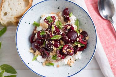 Overhead view of sweet-sour macerated cherries with Marcona almonds, mint, and ricotta, served in as white bowl with blue specks..
