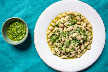 Overhead view of garlic scape pesto in a small bowl next to a wide rimmed, shallow bowl of cooked white beans and pesto