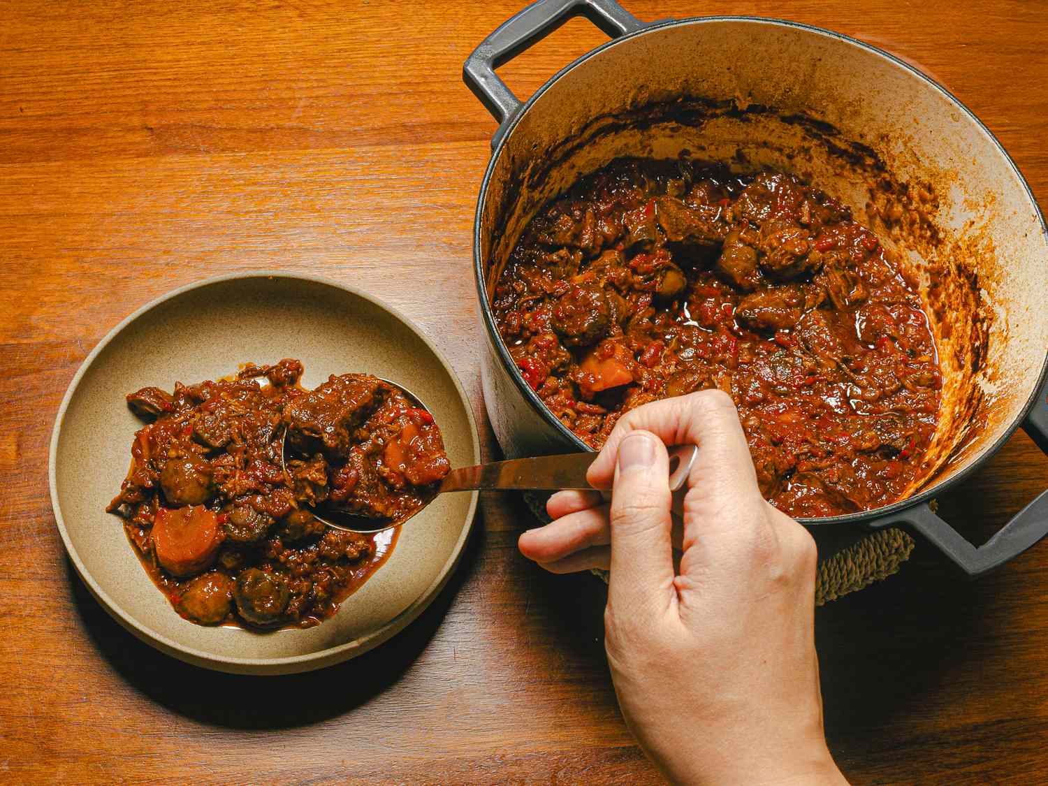 A hand serving Spanish beef stew from a pot into a bowl using a spoon