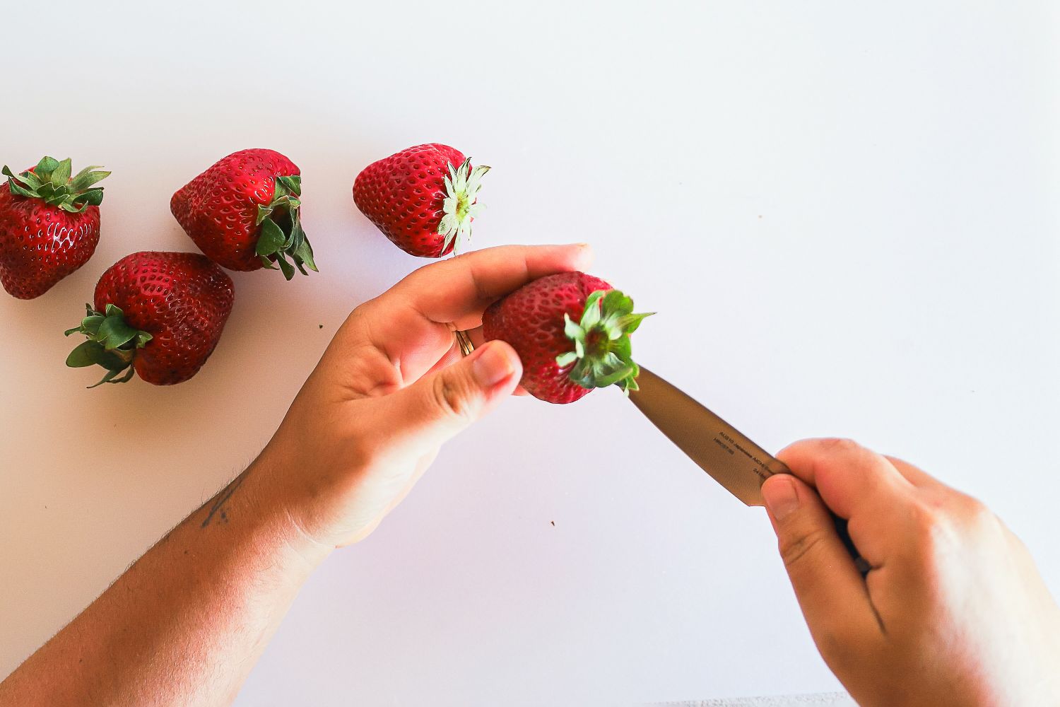 Hands using a Misen knife to hull a strawberry on a white surface with other strawberries nearby