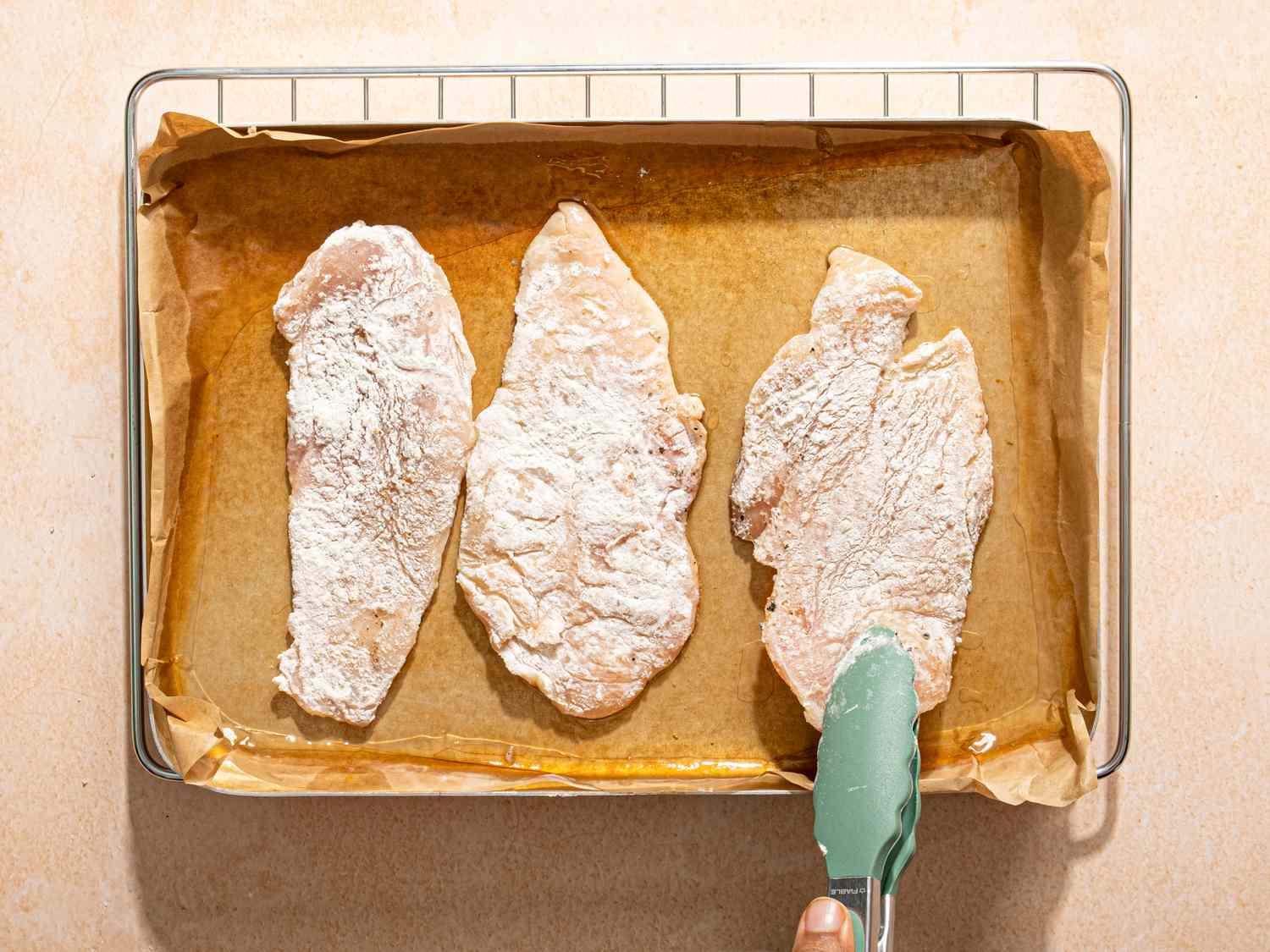 Breaded chicken fillets placed on a baking sheet with a hand holding tongs adjusting one of the pieces