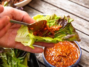 A small bowl of Ssamjang, and someone spooning ssamjang onto a lettuce leaf. 