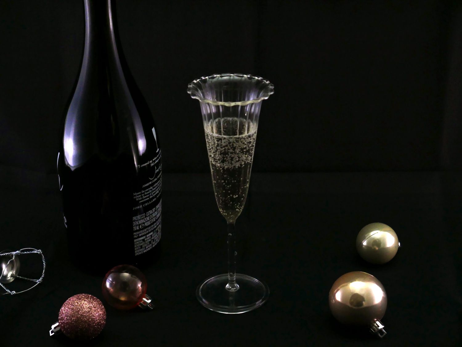 A champagne flute filled with sparkling wine a dark bottle and scattered ornaments on a black background