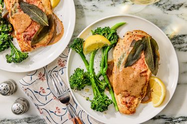 Two plates of chicken saltimbocca with sauce broccolini and lemon wedges on a table setting