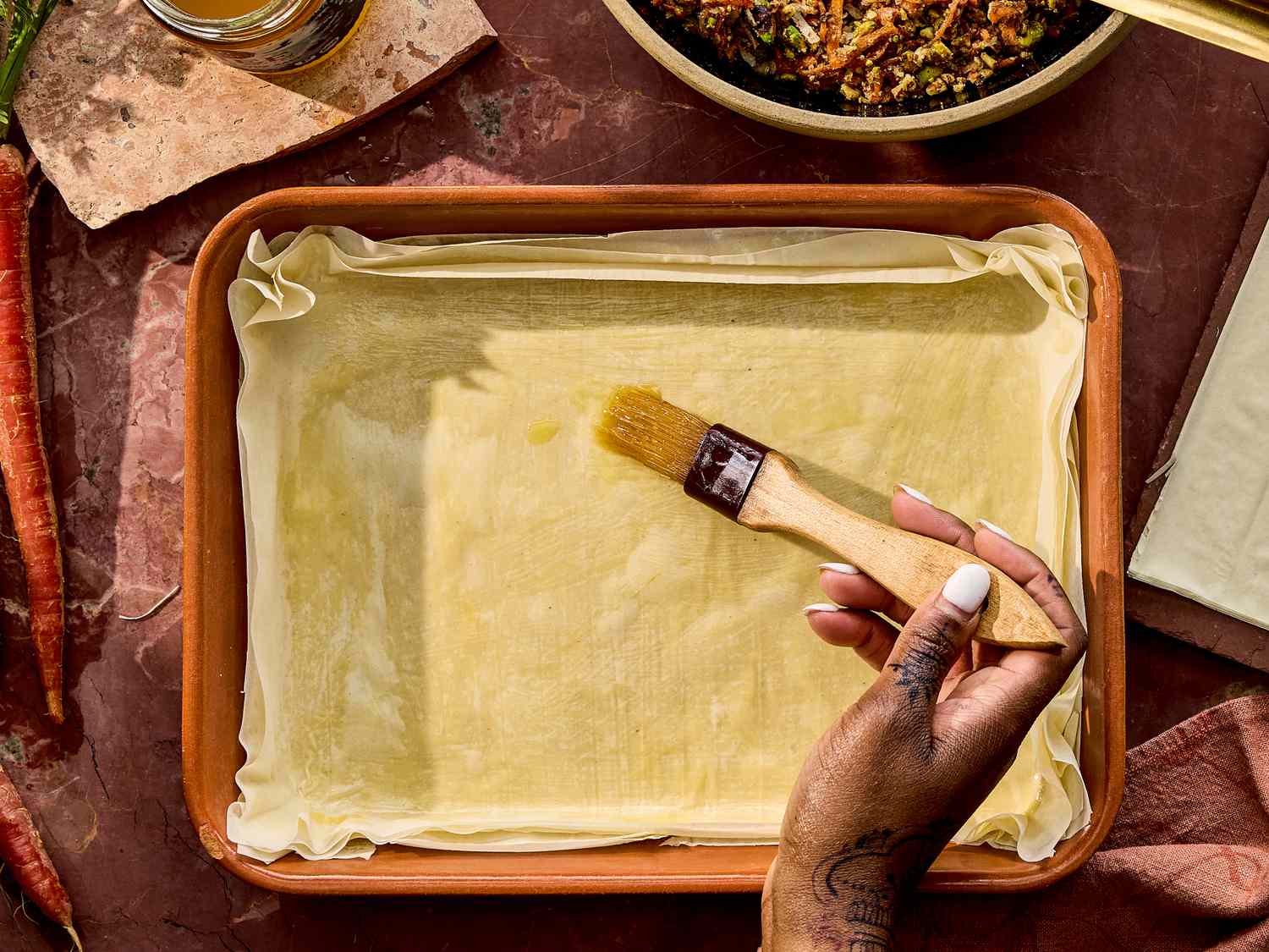 A person's hand brushing phyllo dough layered in a baking dish, surrounded by ingredients and utensils