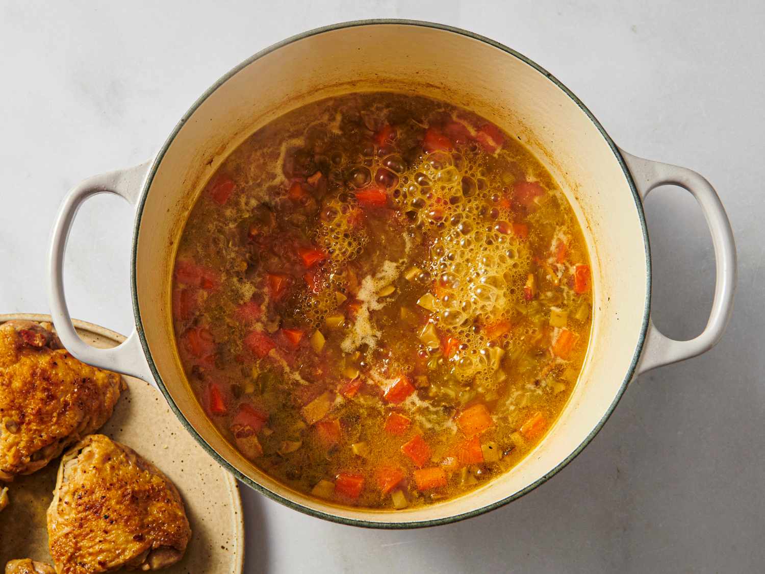 A pot of cooking chicken stew with visible vegetables next to a plate holding cooked chicken pieces