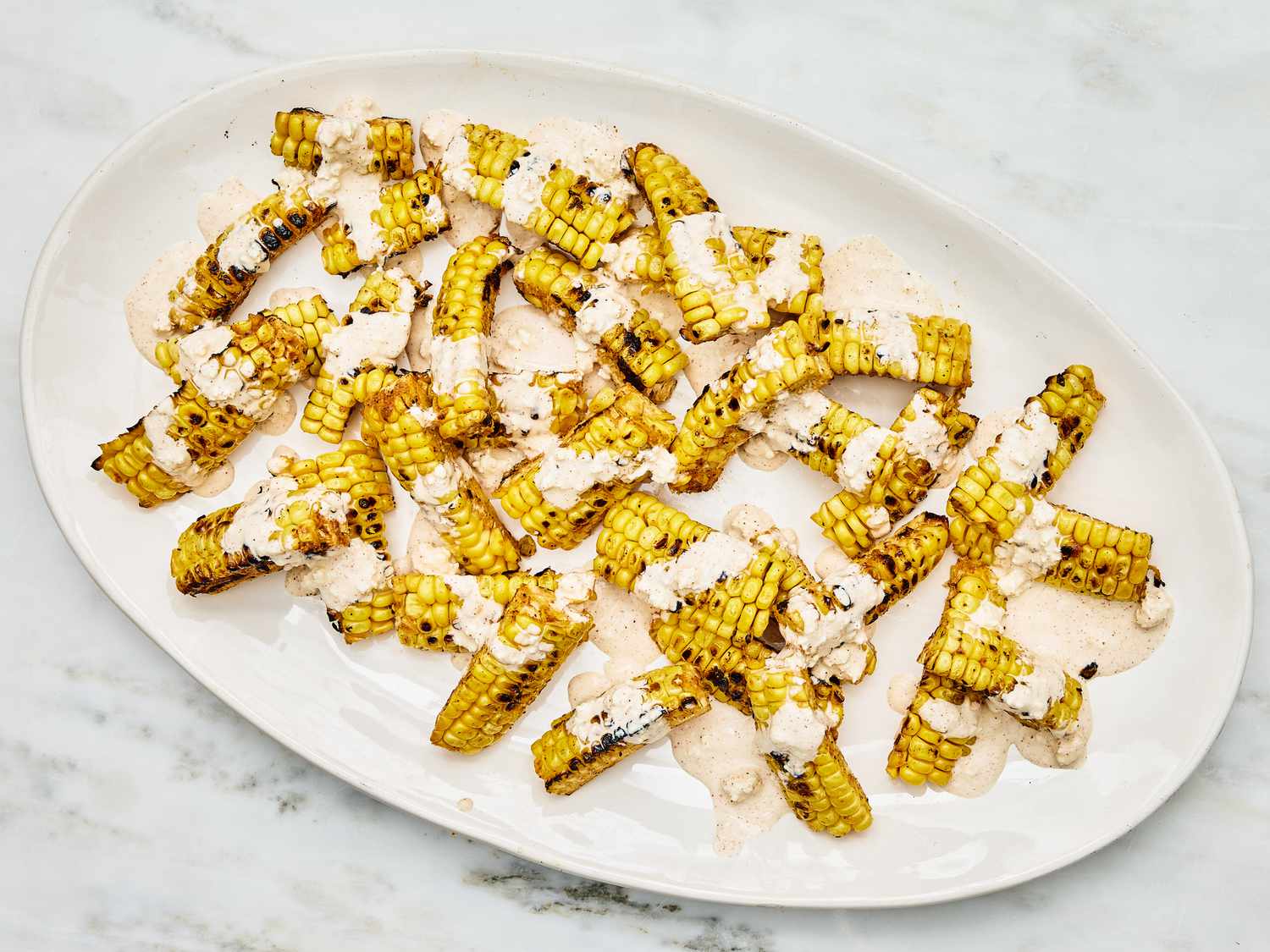 Overhead of thinly sliced grilled corn with cheese sauce on a white dish on a white marble surface