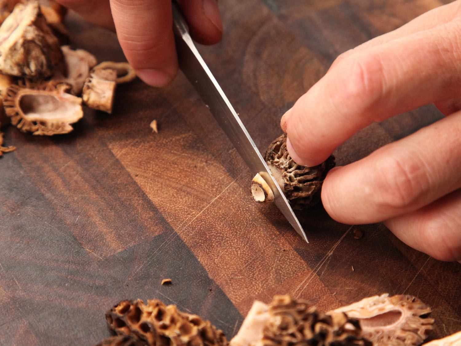 Hands using a knife to slice the tough end off a morel mushroom.