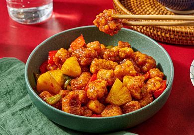 A bowl of Chinese sweet and sour pork with pineapple and vegetables chopsticks holding a piece above the dish