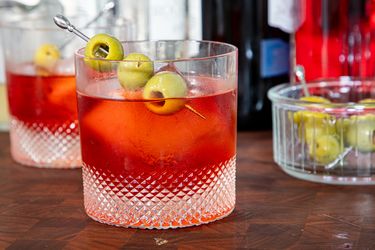 A glass of a cocktail garnished with green olives on a pick placed on a wooden surface with bottles and a dish of olives in the background