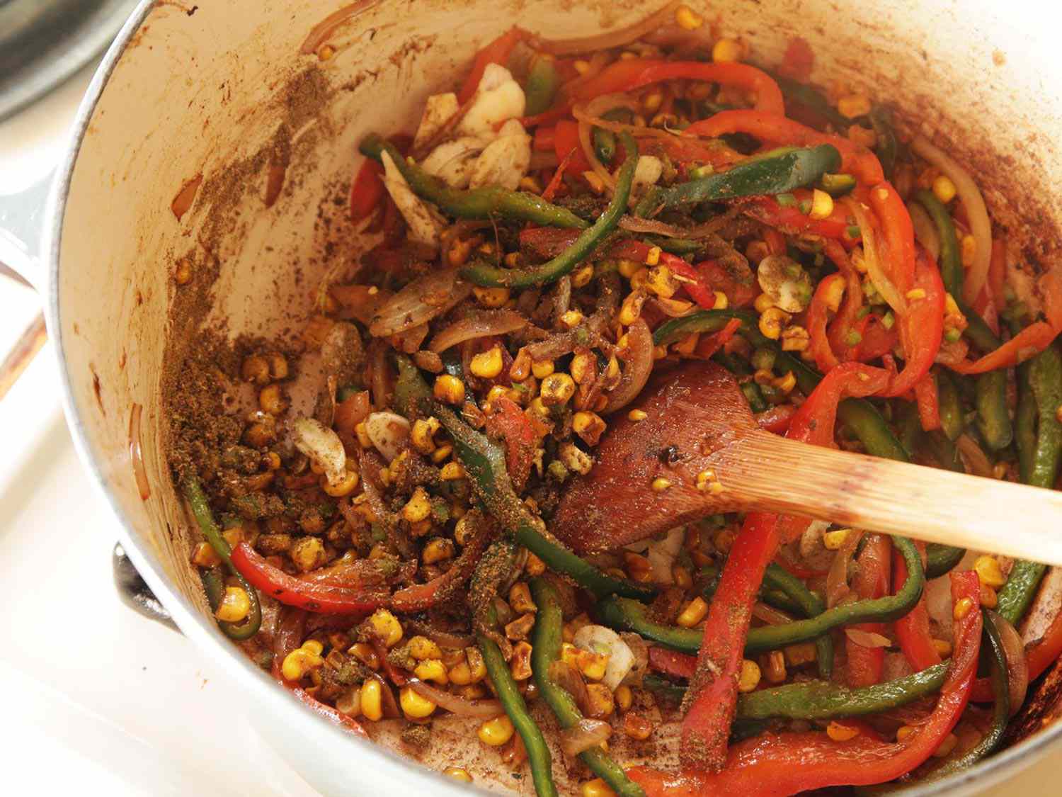 Wooden spoon stirring sliced peppers, corn, and spices together in Dutch oven