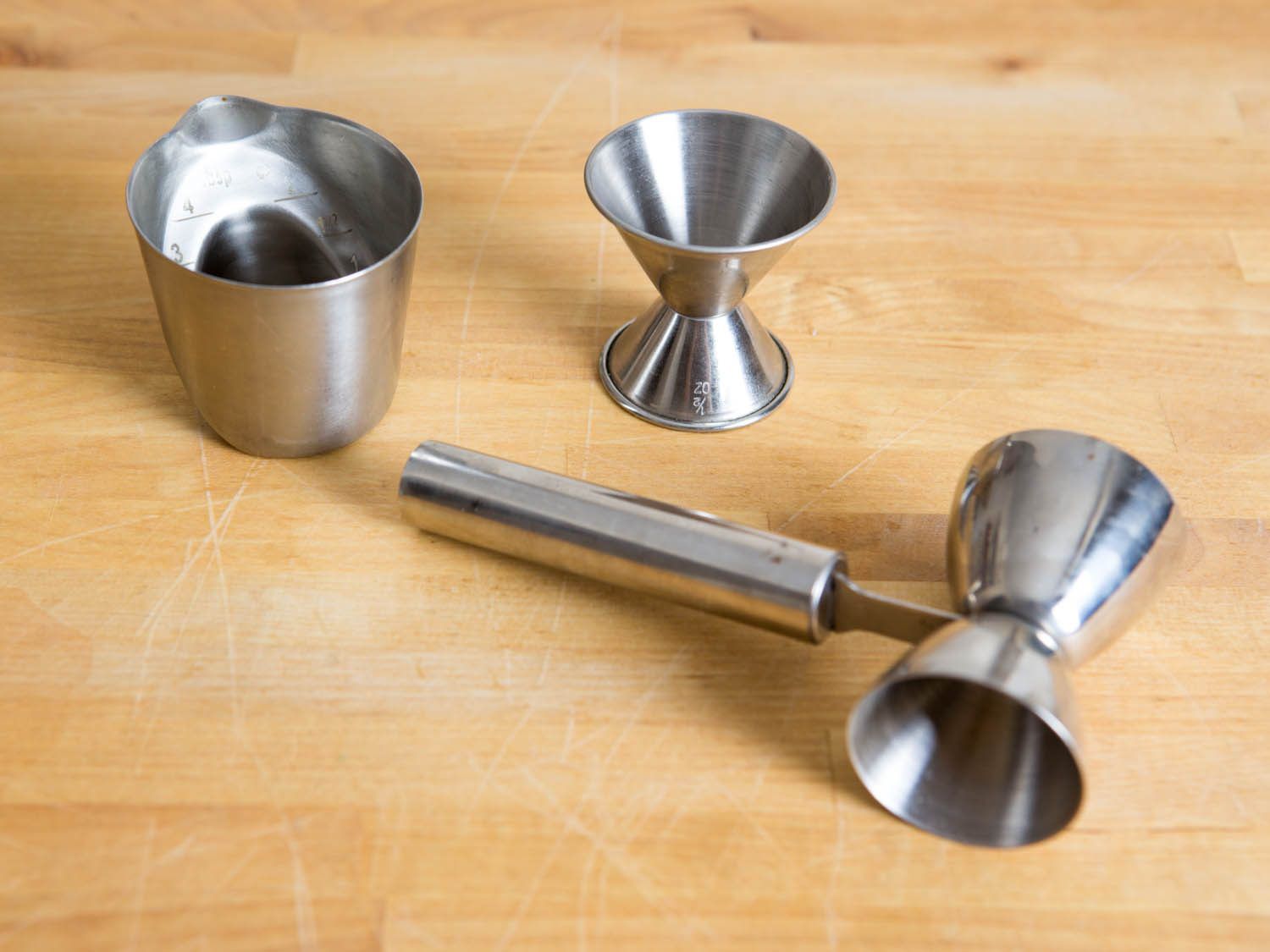 Several styles of jiggers for making cocktails arrayed on a wooden surface.