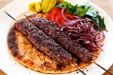 Two Adana kebabs on a plate with flatbread, sumac onions, parsley, tomatoes, and pickled peppers. 