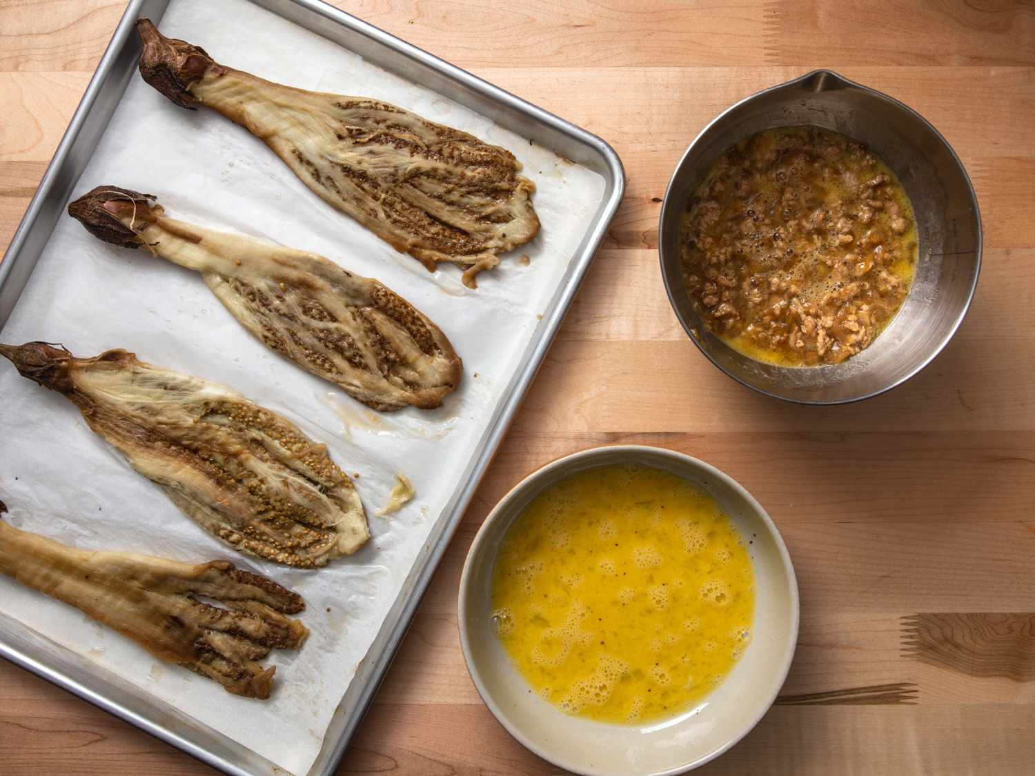 prepared eggplant on a baking sheet with ground pork mixed with egg in one bowl, and beaten eggs in another bowl